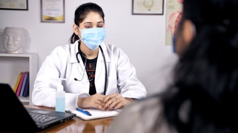 A female doctor wearing a face mask talking to her patient in the clinic. Medium shot of a young Indian medical practitioner in a white uniform prescribing medicines during Coronavirus outbreak