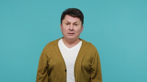 Extremely scared terrified middle aged brunette man screaming holding out his hands, trying to hide and defend himself, panic and frustration. Indoor studio shot isolated on blue background