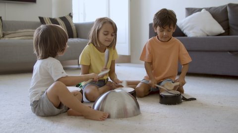 Happy little kids playing in living room with utensils. Blonde girl and two boys smiling, sitting on floor together and knocking on pans. Static camera. Childhood, noise and home activity concept