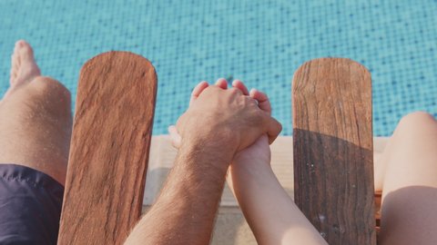 Close up rear view of couple on vacation holding hands as they lie on loungers and sunbathe by the side of outdoor pool shot from behind over their shoulders -shot in slow motion