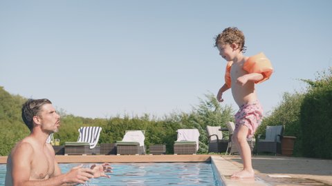 Father catching son wearing armbands as her jumps from side of outdoor swimming pool into water - shot in slow motion