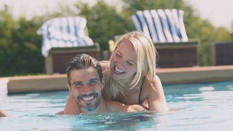 Portrait of loving couple having fun on summer vacation in outdoor swimming pool with man giving woman piggyback in water - shot in slow motion