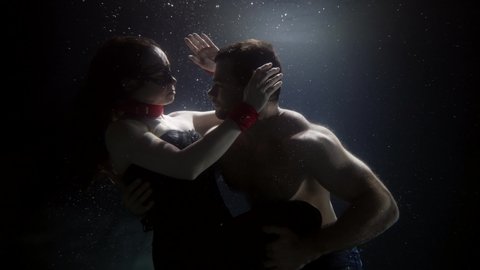 mysterious lovers are hugging and floating underwater, slow motion shot, woman in bdsm suit and half-naked man, embrace and passion