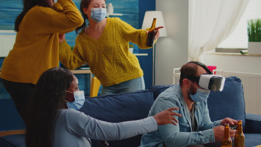 Multiethnic friends enjoying vr tehnology playing video games in living room wearing mask to prevent infection with coronavirus keeping social distancing. Diverse people having fun at new normal party