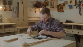 curly joiner master using smartphone  checks measurements , handsome Caucasian young man carpenter in apron and caged shirt holding mobile device sitting at work-desk - Powered by Shutterstock - Get 15% off with code: PIKWIZARD15