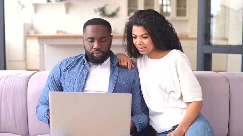 Cheeful African-American couple spends leisure time online with a laptop at home, a charming woman and handsome man sit on the couch, watching movies or shopping online together | Shutterstock HD Video #1069471549