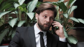 Handsome businessman with a beard in a classic suit is negotiating with partners on the phone in a modern office building on a background of green plants. Business. Social Networking. People. Apps.
