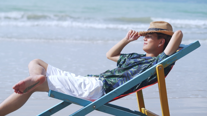 4K Young Asian man tourist resting on sun bed by the sea. Handsome male in beach clothing on beach chair relax and enjoy outdoor lifestyle at tropical island sand beach in summer holiday vacation