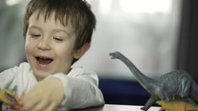Happy little boy playing. The child heals with dinosaur toys on the table and smiles. 4K slow motion beautiful video of home life.