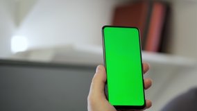 Handheld Camera: Point of View of Man at Modern Room Sitting on a Chair Using Phone With Green Mock-up Screen Chroma Key Surfing Internet Watching Content Videos Blogs Tapping on Center Screen