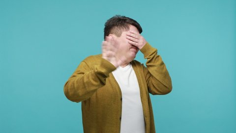 Don't want to look at this. Mature dark haired man in t-shirt and cardigan closing eyes with hand showing stop gesture, confused shy scared to watch. Indoor studio shot isolated on blue background