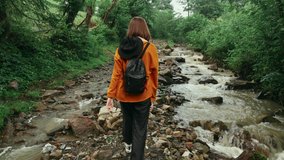 Woman hipster in casual clothes on a hike in the mountains walks along the fireplace stream, view from the back. Travel to mountains concept.