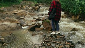Woman tourist in a red shirt walks on a mountain stream, jumps over rocks in a hike. Hipster woman with a bag in her hands on a hike near a mountain river