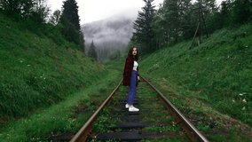 Beautiful hipster woman walking in the evening forest in the mountains, walking on the railway track with a handle bag on his shoulder, view of the back