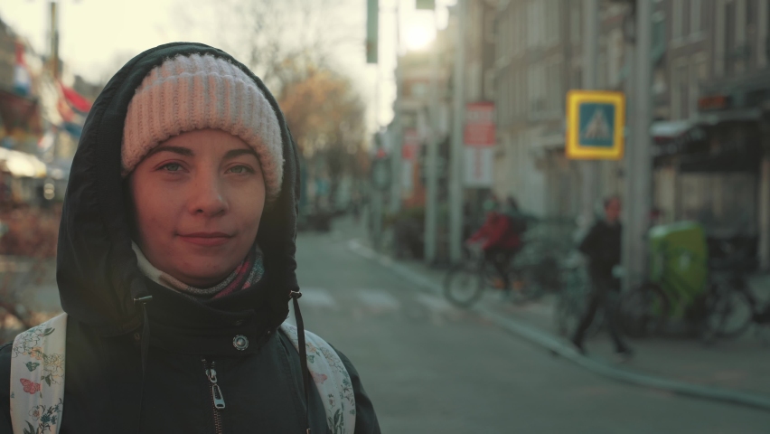 Portrait of cute girl in hat and hood with backpack on blurred background of street in historic center of Amsterdam