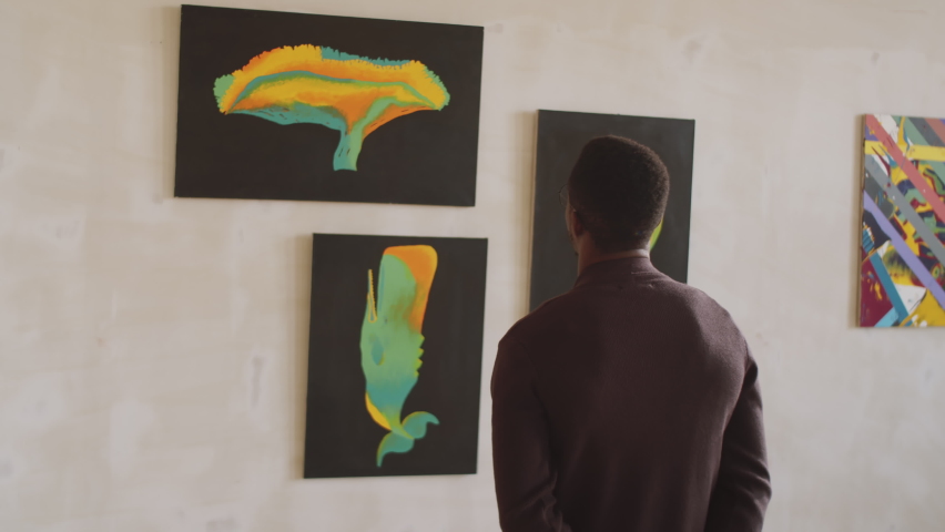 Waist up arc shot of young black male visitor standing in art gallery and looking at abstract paintings on the wall