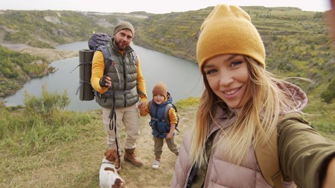 POV shot of happy young woman holding camera and taking selfie or filming herself and her cheerful husband, 5-year-old son and cute Jack Russell Terrier dog while hiking near quarry lake