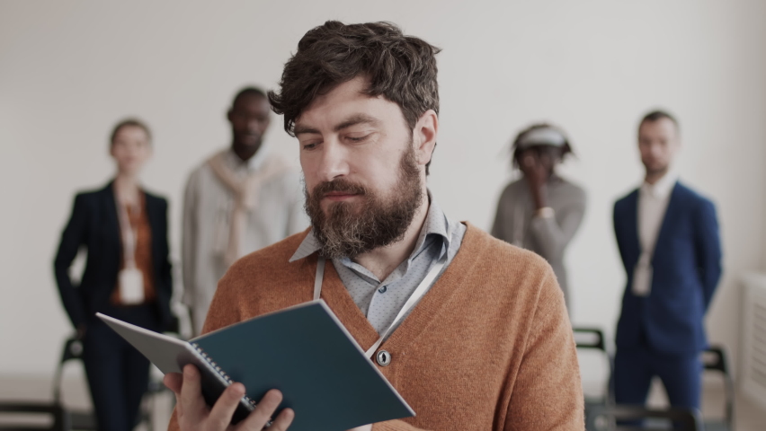 Medium close-up POV of bearded male Caucasian office worker wearing shirt and brown cardigan, reading booklet, then smiling and looking on camera