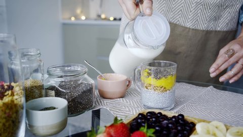 Woman pours fresh gluten free milk into transparent mug with fruits and chia seeds cooking breakfast on table in light kitchen at home close view