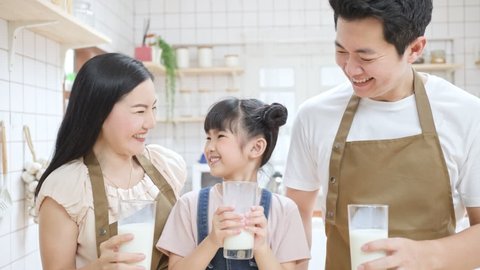 Portrait of Asian happy family clinking a cup of milk and drinking together in kitchen at home. Little kid daughter with father and mother smiling, looking at each other, enjoying spend time in house.