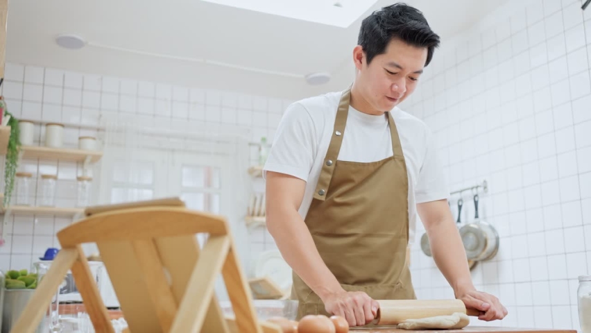 Asian attractive man stay at home in kitchen and practice baking bakery. Handsome father kneads yeast dough with his hands, learn how to make bread by using tablet to look for recipes and ingredients.