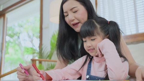 Asian loving mother playing with little kid daughter having fun spending free leisure time painting on paper. Mom teaching young preschool girl drawing picture with color pencil in living room at home