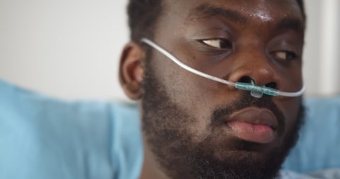 Close up of young african man patient with nasal cannula resting in bed at hospital. Head shot of afro-american sick young man with oxygen tube lying in bed at hospital ward feeling unwell