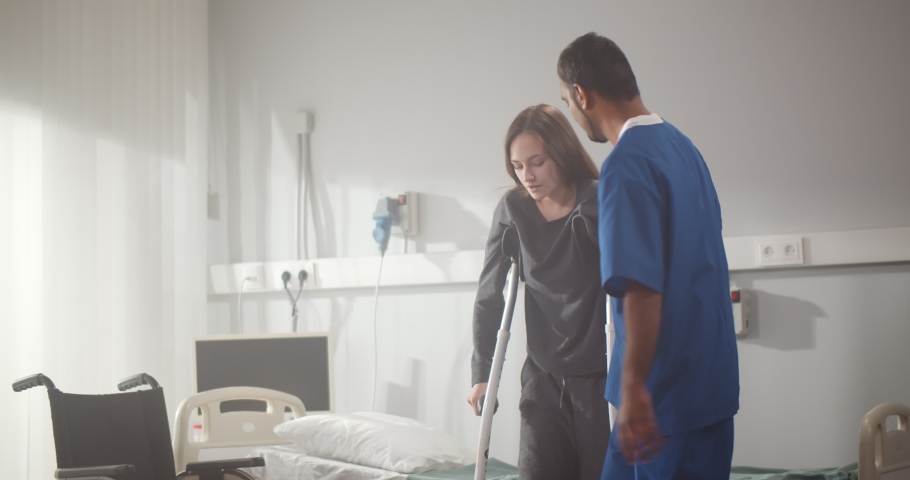 Indian doctor taking care of young female patient in crutch at hospital. Male nurse helping injured woman walking with crutches during rehabilitation treatment in clinic Royalty-Free Stock Footage #1069523182