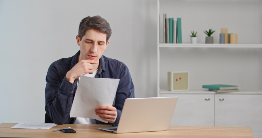 Concentrated caucasian millennial businessman sitting at home desk or in office, looking at documents, reading letter paper, looking in financial statement, receiving notice from bank, doing paperwork Royalty-Free Stock Footage #1069523944