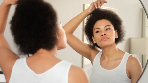 African american woman applies hair care balm styling cosmetics, makes hairstyle in front of the mirror at home.