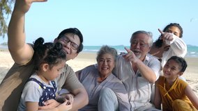 Happy Multi-generation Asian family on summer holiday vacation. Grandparents, parents with two child girl kid enjoy and having fun using smartphone selfie or talking video call together on the beach.