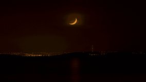 Young moon crescent setting over the horizon, Cardiff industrial skyline timelapse video, dead of the night. Light misty clouds, atmospheric fluctuations distorting the shape of the moon. Dark sea. 
