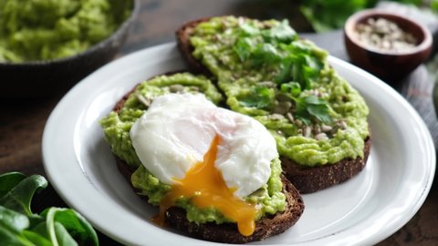 Avocado toast with poached egg. Cutting poached egg with runny egg yolk over rye bread toast with mashed avocado spread. Healthy breakfast or lunch food.