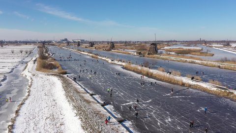 Drone flight over classic Winter landscape in the Netherlands. People ice skate on frozen canals past windmills, enjoying a traditional Dutch sport (a welcome break from Covid-19 lockdown)
