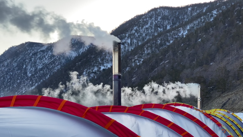 White smoke pours from the chimneys of two stoves of a large winter tent against the backdrop of the hills and forests of Olkhon Island. Frosty Lake Baikal, winter hike. Close-up.