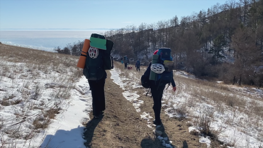 A group of tourists with backpacks walks along the trail to the frozen Lake Baikal. View from the back. Beautiful winter landscape. Winter hike, Olkhon island.