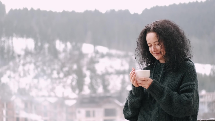 Middle aged woman with curly hair drink coffee on balcony of mountain hotel