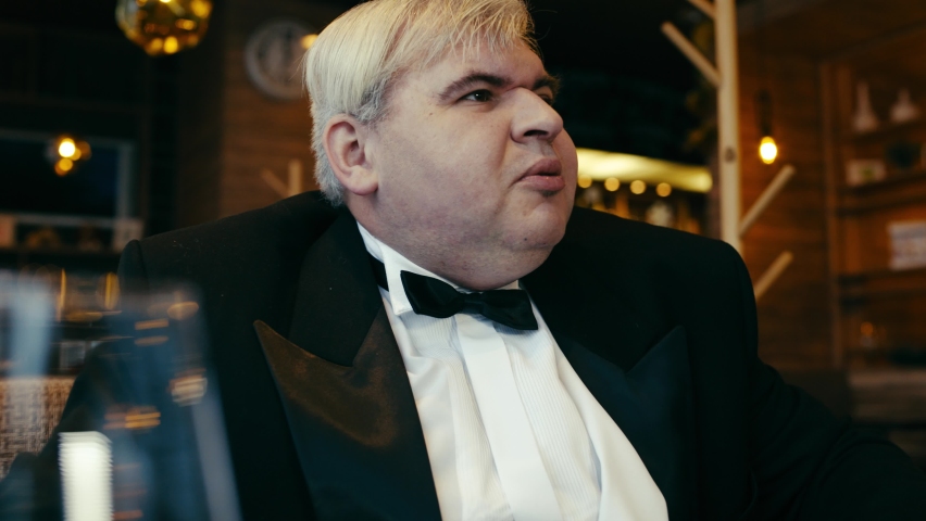 An unpleasant-looking fat man in a tailcoat sits at a table in an expensive restaurant. With a discontented and disdainful expression, he calls the waiter with a gesture of his hand