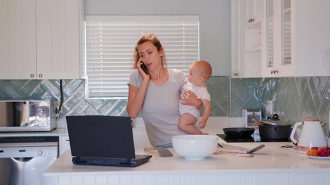 Mother multi-tasking, holding baby infant and using computer laptop at home. Candid authentic and real life mom working and parenting.