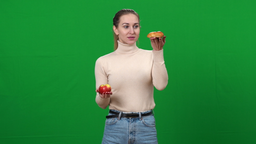 Slim woman choosing healthful apple holding fruit and bun in hands. Portrait of young Caucasian lady selecting food on green screen. Healthy and unhealthy eating concept.
