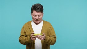 Addicted mature man in white t-shirt and cardigan playing online game on modern smartphone, controlling character in action simulator, leisure activity. Indoor studio shot isolated on blue background