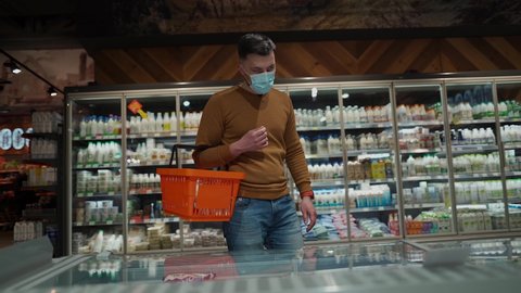 Young caucasian man in face mask choosing dairy products at supermarket during coronavirus pandemic. Male shopping at grocery store wearing facemask, shopping basket in hand. Topic new norm covid 19