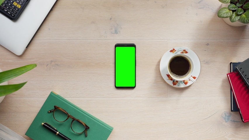Above shot of Male hand picking up cup of coffee next to vertical phone with green screen. Light wood desk, glasses, fountain pen, calculator, books on background. 4k POV close-up flat lay Royalty-Free Stock Footage #1069566073