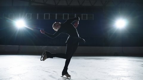 graceful female figure skater is performing spin on ice rink in darkness, moving against bright soffit, silhouette shot of professional sportswoman