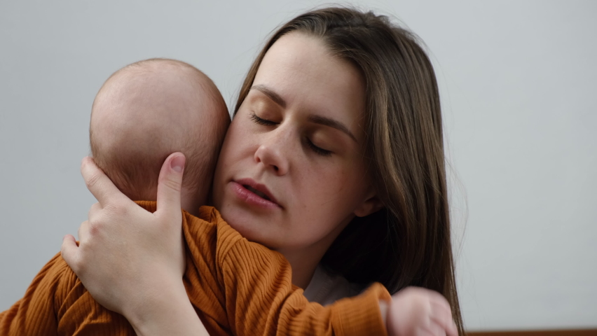 Head shot side view tired caring young mother yawning while holding and cute baby infant falling asleep together sitting on bed at home. Exhausted caucasian mom wants sleeping. Motherhood concept