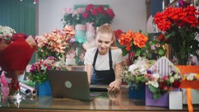 Female florist discusses the order by talking on video call, using a laptop and screen tablet in a flower shop, working online, the female chooses flowers for the customer.