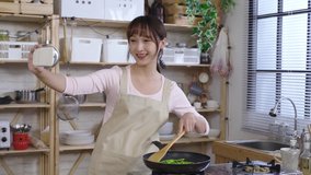 chinese lady making lunch by the gas cooker and holding a smartphone is shaking her head with a smile while video chatting with her friend at home.