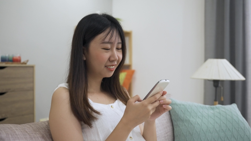 Portrait beautiful asian girl is chuckling while enjoying funny social media feeds on her smartphone in the bright living room at home. | Shutterstock HD Video #1069597954