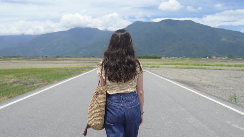 rear view asian lady is pushing back hair while walking ahead in the center of Mr. Brown Avenue with large open farmland on sides and hills in the end.