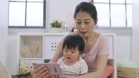 asian lovely baby is smiling at the screen with innocence as her cheerful mother is chatting on the phone during a video call in a bright home interior.